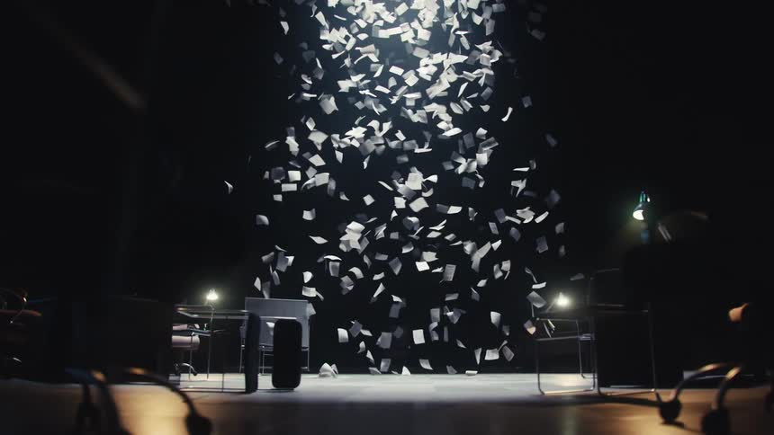 A storm of white papers rains down from a spotlight onto a dark, abandoned office floor in slow motion