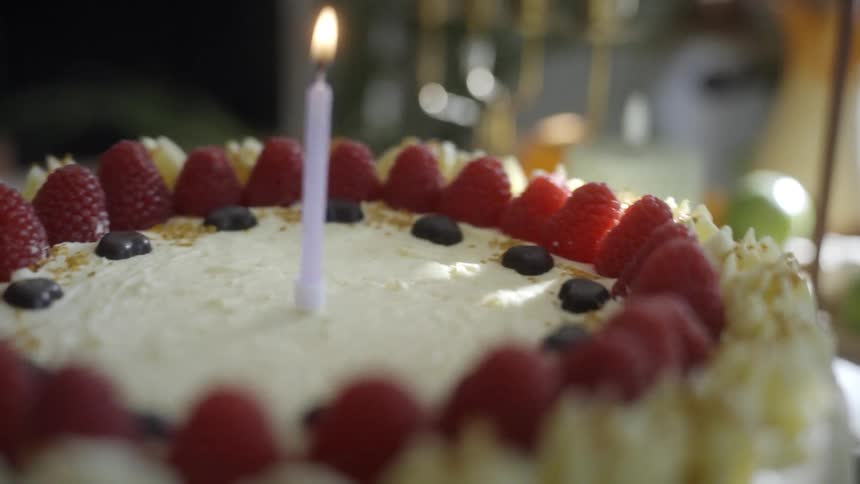 A single lit birthday candle sits in the middle of a raspberry and cream cake as the camera slowly rotates around it