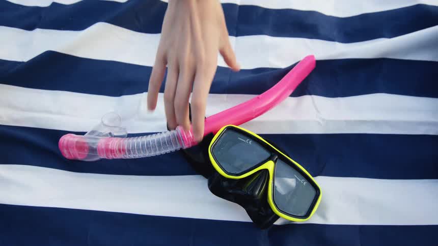A hand picks up a neon snorkel and mask from a blue and white striped beach towel in slow motion