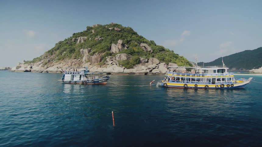 Two tour boats are anchored in a tranquil blue bay near a lush, rocky island as a person jumps into the water