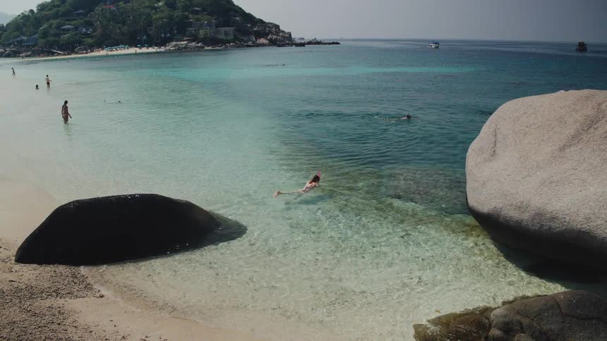 A woman with snorkel gear swims in the clear shallow water of a tropical beach with large boulders