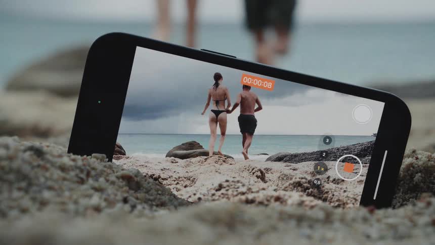 Creative shot of a smartphone propped up in the sand recording a couple holding hands and running towards the ocean