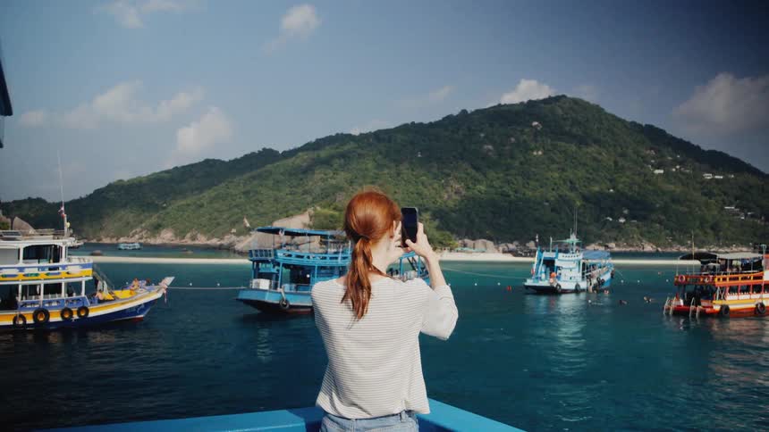 A female tourist stands on a boat and takes a picture with her smartphone of a stunning tropical island bay