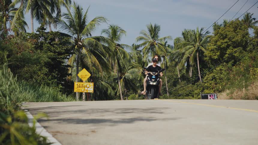 A young couple enjoys a scooter ride down a paved road surrounded by lush tropical palm trees on a sunny day in Thailand