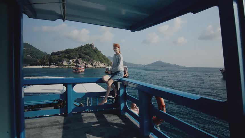 A young barefoot woman sits on the railing of a moving tour boat, smiling as she enjoys the view of a tropical island on a sunny day