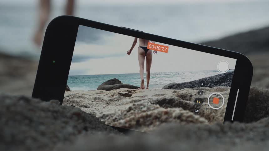 A smartphone screen shows a recording of a woman in a bikini walking barefoot on a sandy beach towards the ocean
