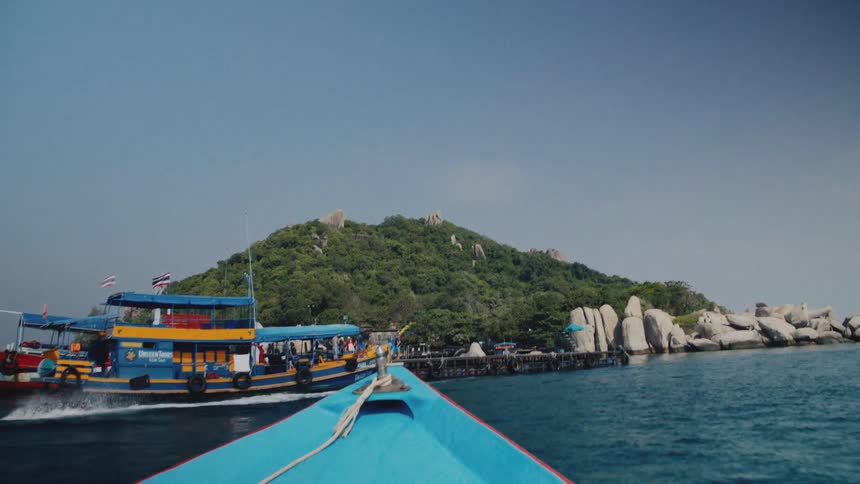 A point-of-view from a boat sailing towards a lush tropical island and pier on a bright sunny day