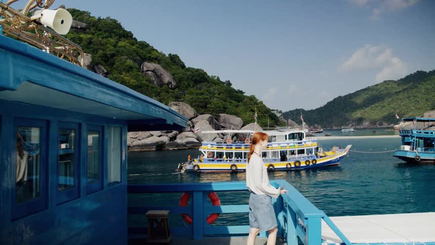 A young woman with red hair walks to the railing of a boat to enjoy the scenic view of a tropical island bay in Thailand