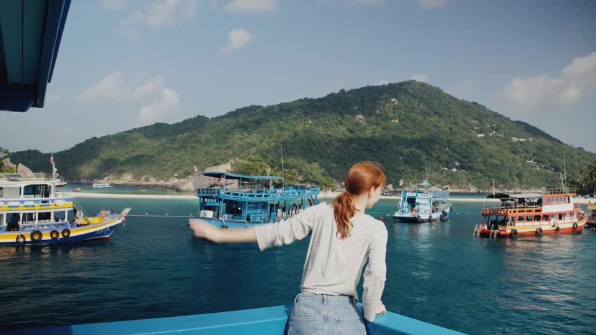 A young woman waves in slow motion from the back of a boat as it departs a scenic tropical island harbor filled with colorful boats
