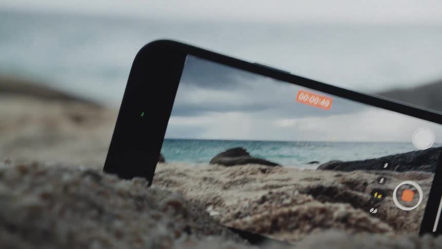 A smartphone sits propped up in the sand, recording a person's feet walking on a cloudy beach by the ocean