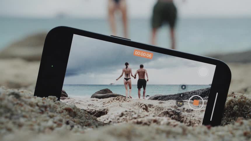 A creative shot shows a smartphone recording a video of a couple running into the ocean on a cloudy beach