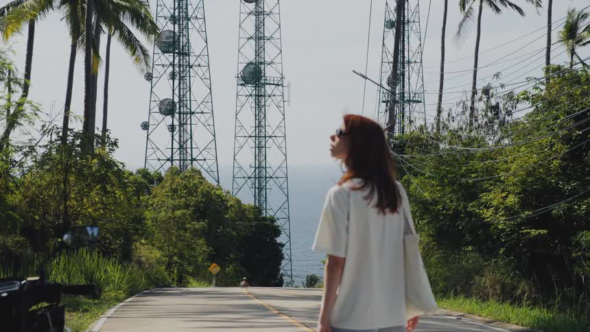 A young woman walks down a tropical road lined with palm trees towards large communication towers overlooking the ocean