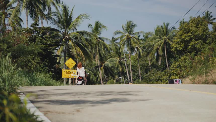 A couple on a motorcycle drives away in slow motion down a scenic tropical road lined with palm trees