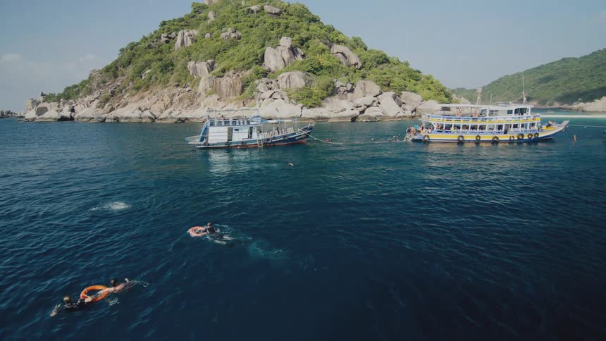 Tourists swim and snorkel in a beautiful blue tropical bay with tour boats anchored near a lush, rocky island