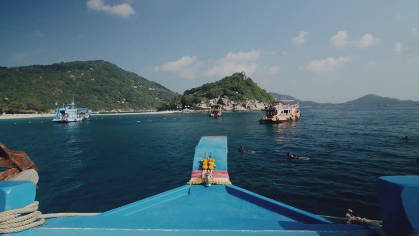 A view from the bow of a blue boat shows other tour boats anchored in a tropical bay as people snorkel in the clear water