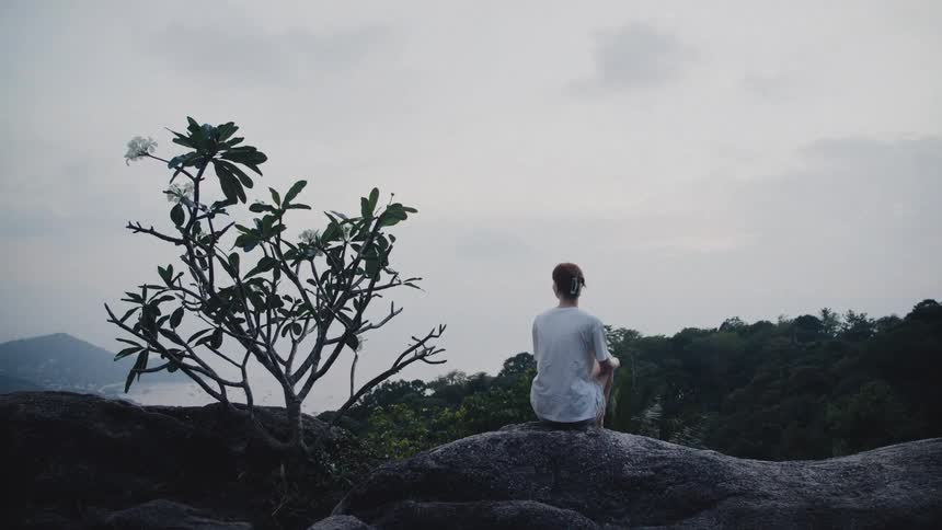 A person sits peacefully on a large rock overlooking a tranquil sea and lush tropical landscape on a cloudy day