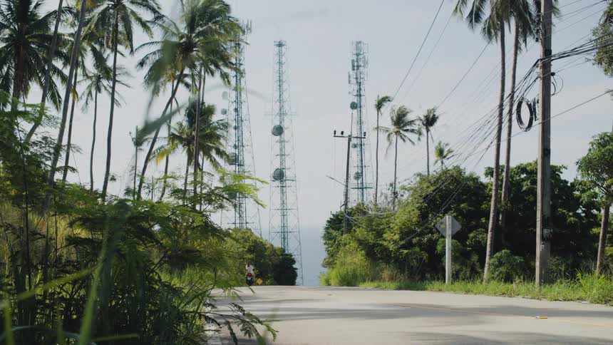 A couple rides a scooter away from the camera down a lush tropical road lined with palm trees and communication towers in the distance