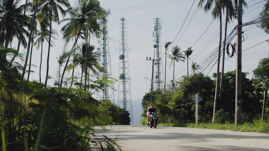 A couple on a scooter drives up a tropical hill road with palm trees and telecommunication towers overlooking the sea