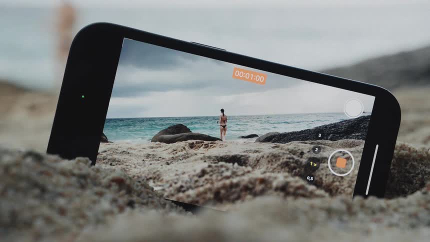 A phone in the sand captures a video of a woman in a bikini walking toward the ocean on a cloudy beach