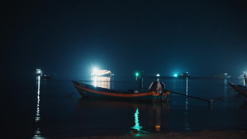 A traditional longtail boat rests in a tranquil harbor at night with brightly lit ships in the distance