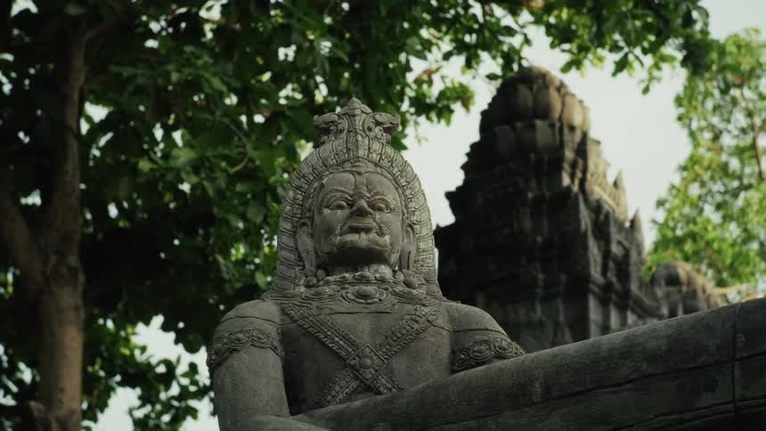 An ancient stone statue of a grimacing guardian figure stands watch at a temple, with lush trees and architecture in the background