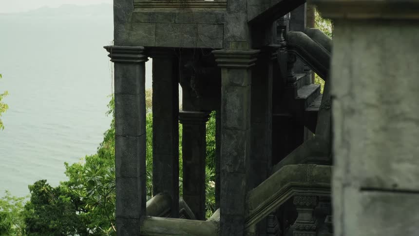 A slow tilt and pan from lush tropical foliage reveals an ancient stone temple overlooking the ocean