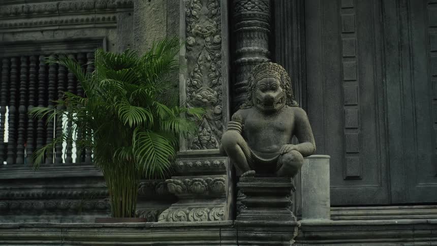 A gray stone statue of a mythical creature guards the ornate entrance to an ancient temple next to a vibrant green palm plant