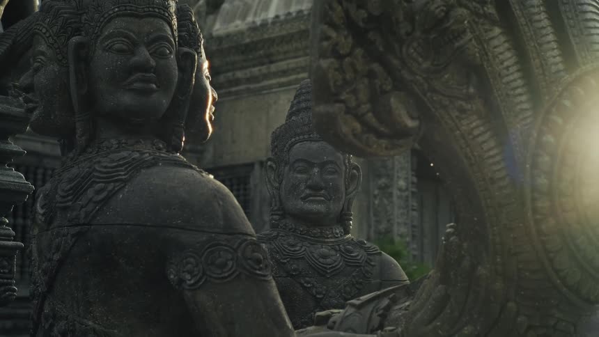 Slow camera movement reveals intricate stone statues of ancient deities as golden sunlight flares behind them