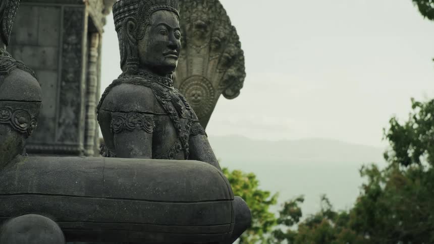 A slow pan up reveals ancient, weathered stone guardian statues at a seaside temple overlooking the ocean