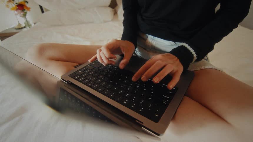 Close up of a woman's hands typing on a laptop keyboard while she sits on a comfortable bed