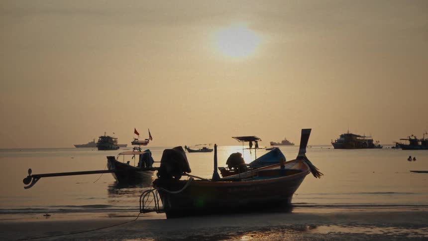 Traditional long-tail boats rest on a tranquil beach at sunset, silhouetted against the golden sky and calm ocean