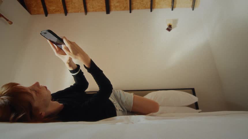 A young woman with red hair relaxes in bed, scrolling and smiling at her smartphone