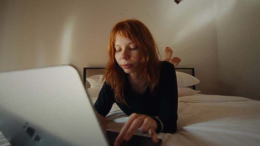 A focused young woman with red hair relaxes in bed while browsing the internet on her laptop computer