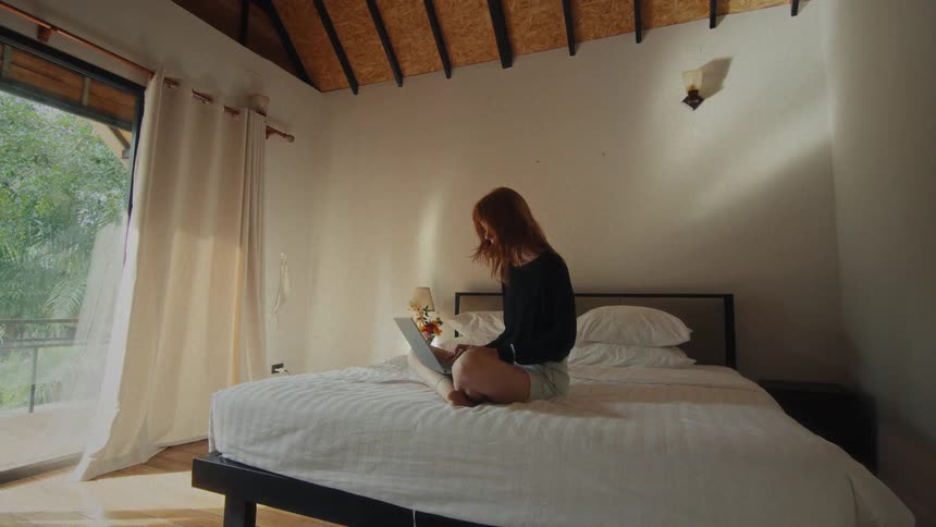 A young woman works remotely on her laptop while sitting cross-legged on a bed in a bright, airy hotel room