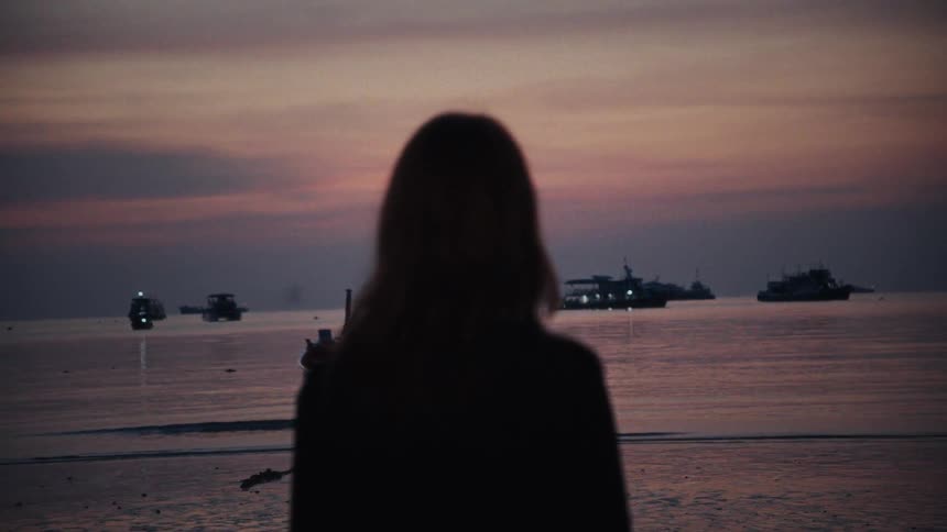 A silhouetted woman stands on the shore watching boats on the ocean as the focus shifts to her during a beautiful twilight
