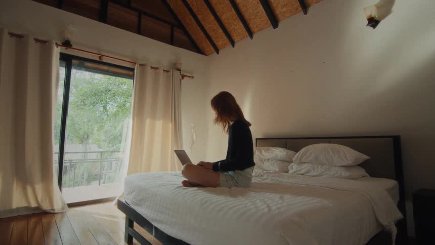 A woman works on her laptop while sitting on a large white bed in a brightly lit bedroom with a balcony view