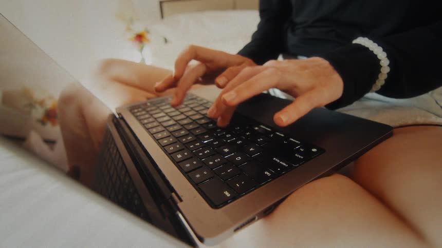 Close-up of a person's hands typing on a laptop keyboard while working from the comfort of their bed