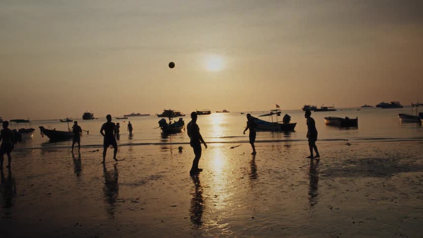 Silhouetted figures enjoy a leisurely game of beach soccer as the sun sets over the water in beautiful slow motion