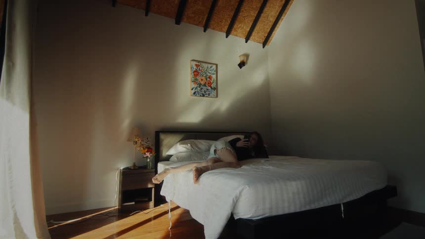 A woman in shorts and a long-sleeve shirt playfully relaxes in bed with her phone in a sunlit bedroom