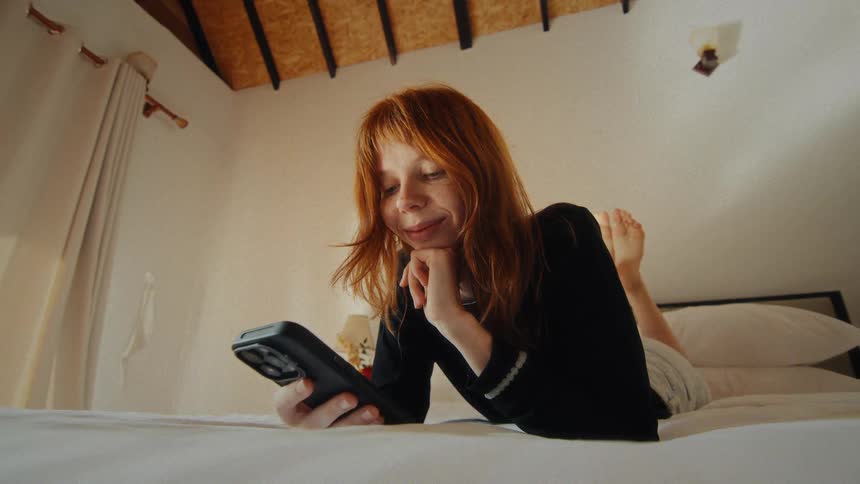 A relaxed, smiling redhead with freckles lies on her stomach in a cozy bedroom, happily using her smartphone