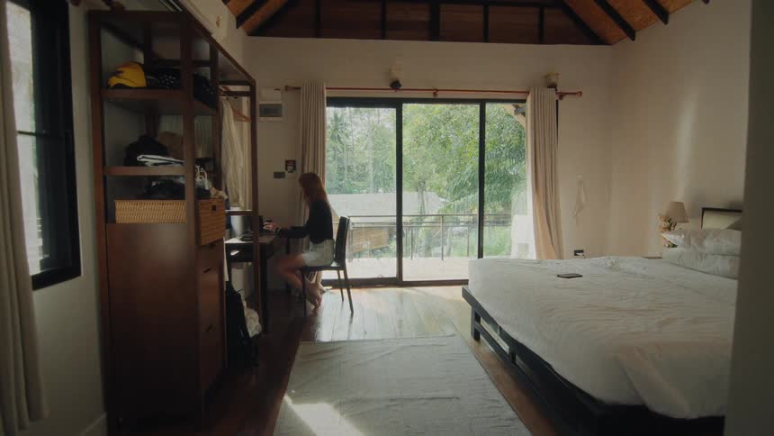 A woman gets up from bed, opens the curtains, works on her laptop, and walks out onto the balcony of her sunlit tropical bedroom