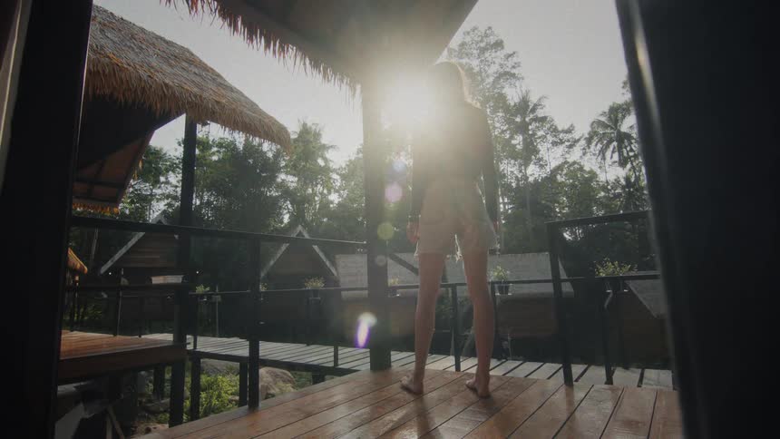 A woman walks out onto the sun-drenched wooden deck of her tropical jungle bungalow to enjoy the serene morning view