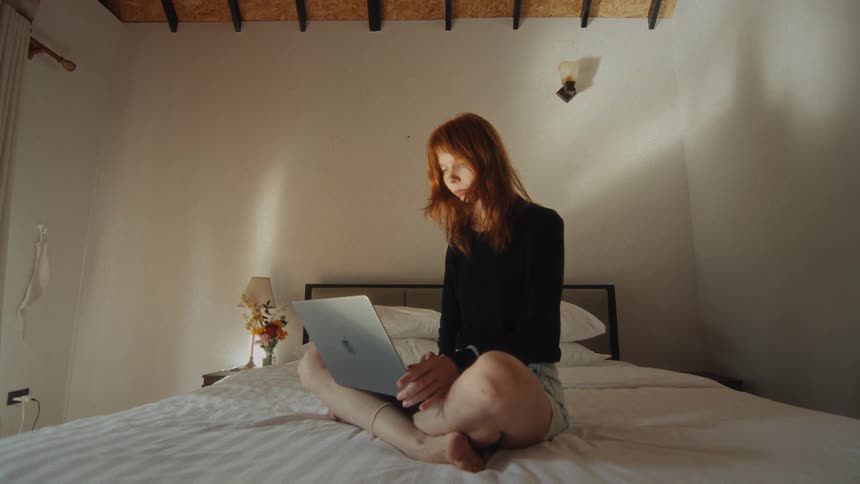 A young redhead woman sits cross-legged on her bed, attentively typing on her laptop in a cozy, modern bedroom