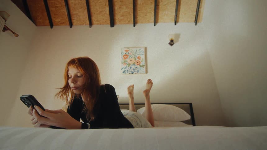 A young woman with red hair relaxes on her bed, scrolling through her phone while lying on her stomach