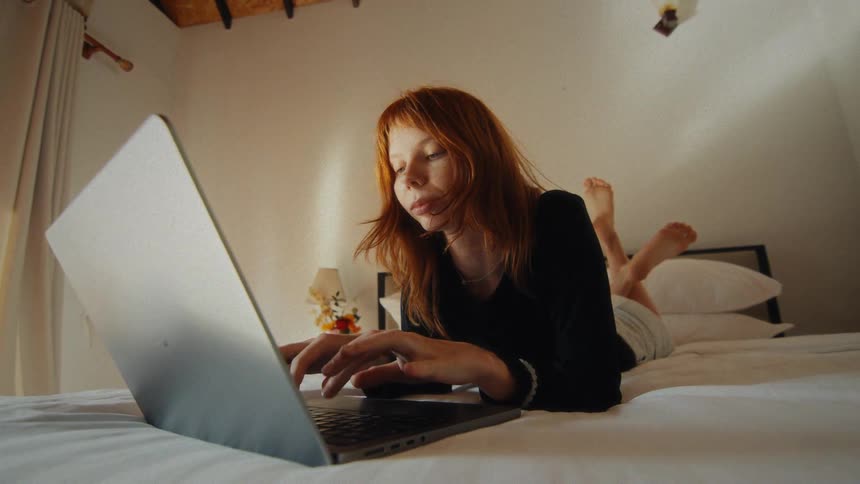 A young woman with red hair lays comfortably on her bed while typing on her laptop in a sunlit bedroom
