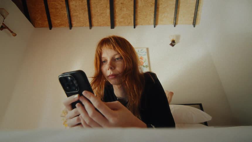 Low angle shot of a young redhead woman lying on her bed and casually scrolling through her smartphone in a bright, cozy bedroom