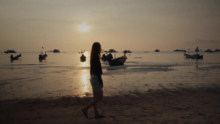 A woman walks in slow motion along a tranquil beach at sunset as traditional longtail boats sit in the calm water