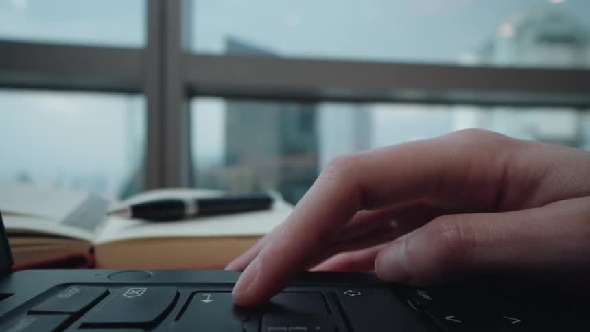 A person's hand scrolls and clicks on a laptop trackpad in an office with a view of the city in the background