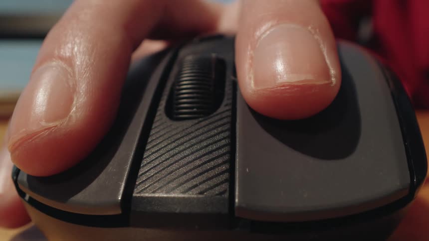 A macro dolly shot pushes in on a person's fingers as they click and scroll a wireless computer mouse on a desk