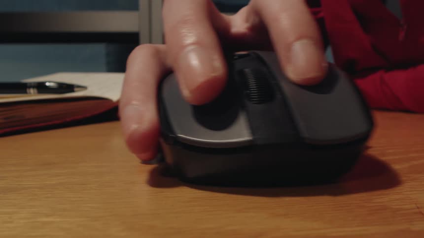 A close-up of a person's hand clicking and moving a wireless computer mouse on a wooden desk